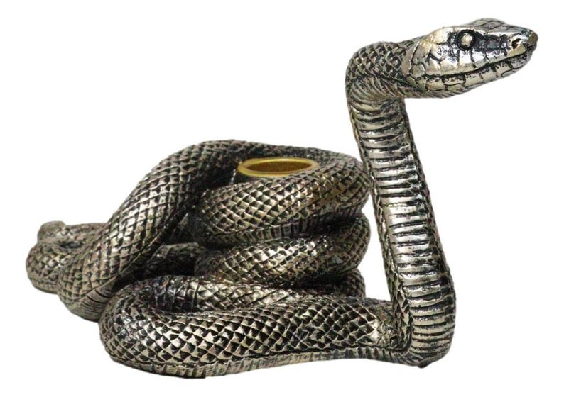K&ouml;nnte beinhalten: Silberfarbene Schlangenfigur mit gewundenem K&ouml;rper und aufrechtem Kopf. Die Schlange hat detaillierte Schuppen und einen goldfarbenen Ring in der Mitte, wahrscheinlich zum Halten einer Kerze. Die Figur ist ein Dekorationsartikel.