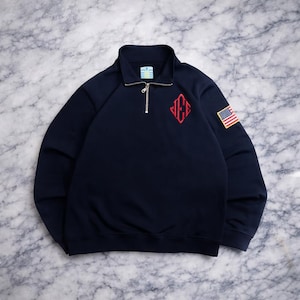 May include: Navy blue sweatshirt with a quarter-zip collar. Features a red embroidered logo on the chest and an American flag patch on the sleeve. The sweatshirt has long sleeves and a ribbed collar.