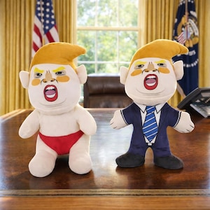 Könnte beinhalten: Zwei Plüschpuppen mit übertriebenen Gesichtszügen und gelben Haaren. Eine Puppe trägt einen roten Windel, die andere einen marineblauen Anzug und eine blaue Krawatte. Beide Puppen haben den Mund offen und stehen auf einem Holztisch.