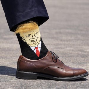 Peut inclure: Une chaussette noire avec un dessin de caricature d'un homme en costume avec une cravate rouge et une coiffure blonde. La chaussette est portée avec une chaussure en cuir marron. La personne porte un pantalon bleu foncé.