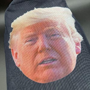 May include: Black mesh face covering featuring a full-face image of a man with fair skin and blond hair. The image is printed on the front of the covering, which is made of a dark, semi-transparent material.