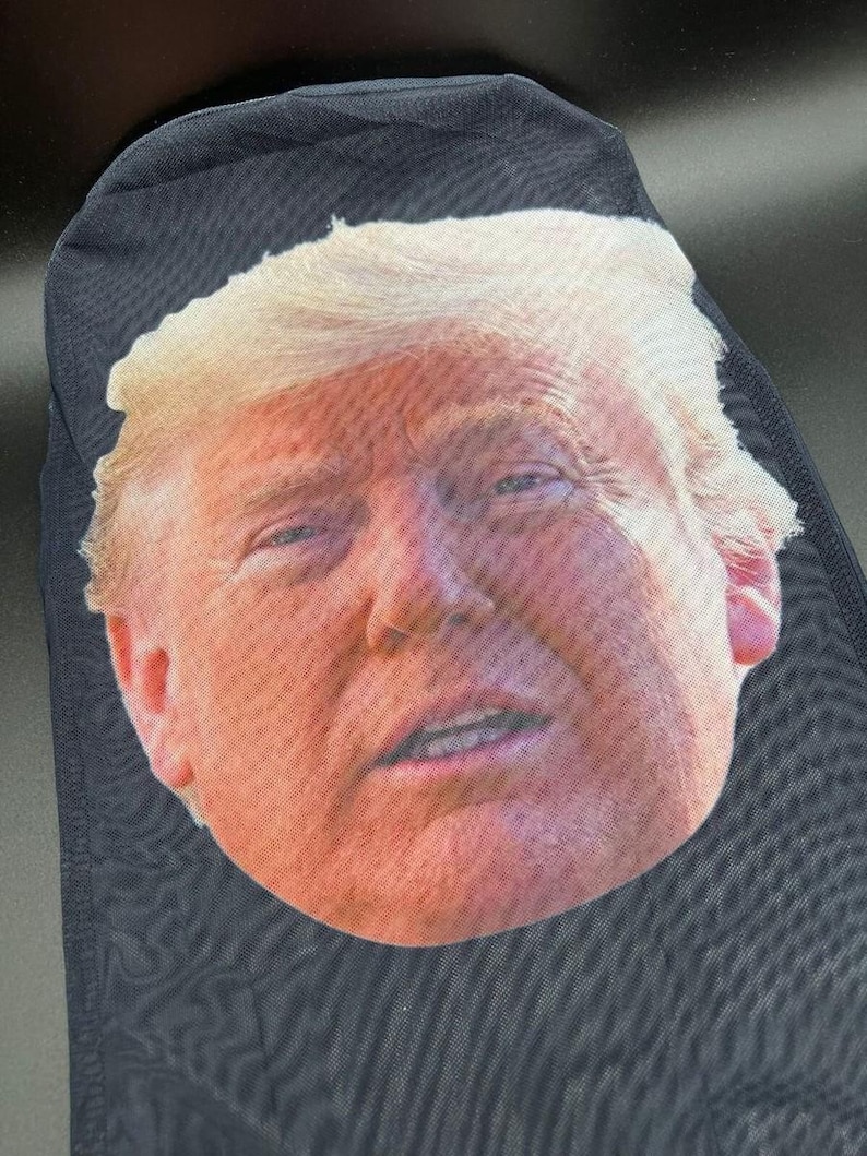 May include: Black mesh face covering featuring a full-face image of a man with fair skin and blond hair. The image is printed on the front of the covering, which is made of a dark, semi-transparent material.