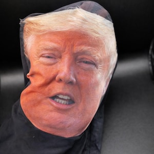 May include: A black face covering featuring a printed image of a person's face with fair skin and blonde hair. The face has a slightly distorted appearance.