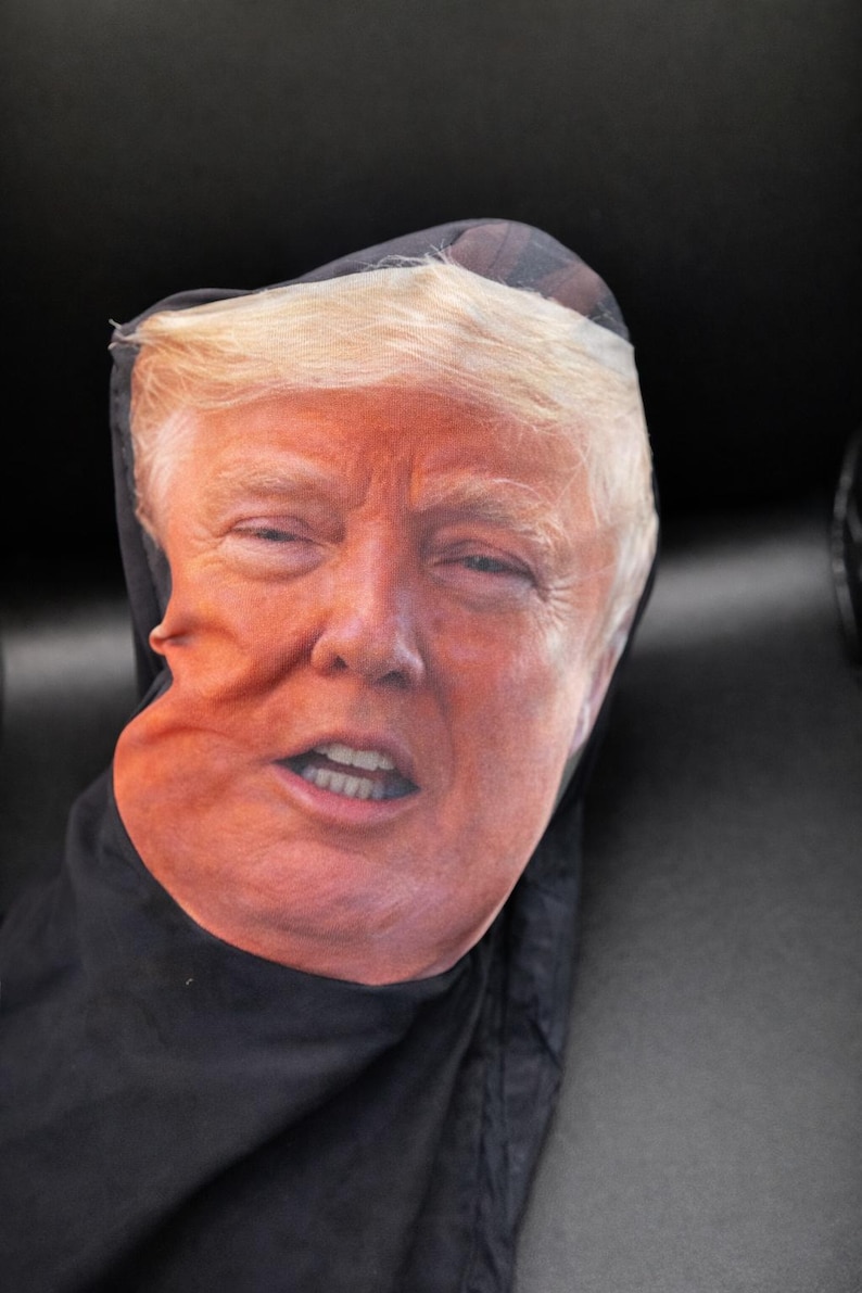 May include: A black face covering featuring a printed image of a person's face with fair skin and blonde hair. The face has a slightly distorted appearance.