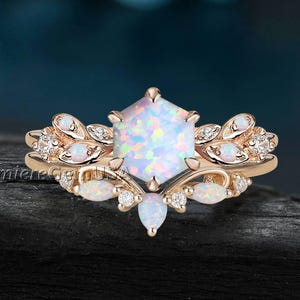 May include: Rose gold ring with a large, hexagonal opal center stone. The ring features smaller opals and clear accent stones, with a leaf-like design on the band. The ring is set against a dark background.