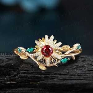 May include: A gold-toned ring featuring a central red gemstone surrounded by a flower design with white accents. The ring has a floral and leaf motif with green and blue gemstones. The ring is set on a dark surface.