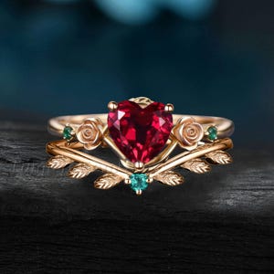 May include: A gold-colored ring set featuring a heart-shaped, deep red gemstone as the centerpiece. The ring is adorned with rose and leaf details, and accented with small green gemstones. The set includes a matching band with leaf accents.