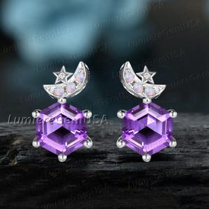 May include: A pair of silver earrings featuring a geometric purple amethyst gemstone. Each earring has a crescent moon and star design with small, round, opalescent stones. The earrings are set against a blurred background.