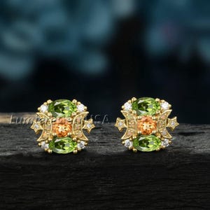 May include: A pair of gold stud earrings. Each earring features two oval-cut green gemstones, a central orange gemstone, and small white gemstones. The earrings have a decorative gold frame with star-like accents. The text "Lumiere Gem USA" is visible.