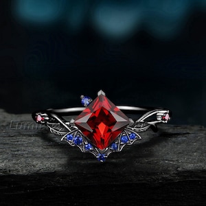 May include: A black ring with a large, square, red gemstone as the centerpiece. The ring is adorned with smaller blue and red gemstones, and features a decorative wing-like design. The ring is set against a dark background.