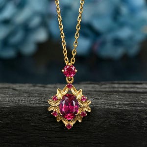 May include: A gold necklace featuring a central oval-cut deep pink gemstone surrounded by a decorative metal frame with small round pink stones. A single round pink stone sits above the pendant, connected to a delicate gold chain link necklace.