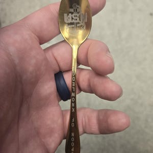 May include: A gold-colored spoon with the text "USN" and an anchor design engraved on the spoon's head. The handle features the words "SPOON OF KNOWLEDGE" vertically. A unique collectible or gift item.