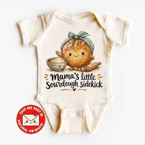 Könnte beinhalten: Ein cremefarbener Kurzarm-Babybody mit einer Grafik eines lächelnden Sauerteiglaibs, der ein blaues gepunktetes Stirnband trägt, neben einer Schüssel und einem Nudelholz. Der Text lautet Mama's little Sourdough sidekick in schwarzer Schrift.