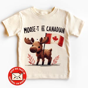 May include: Cream-colored t-shirt featuring a cartoon moose holding a Canadian flag. The moose has brown antlers and fur, with the text "MOOSE-T BE CANADIAN" above it. A red and white sticker with text is at the bottom.
