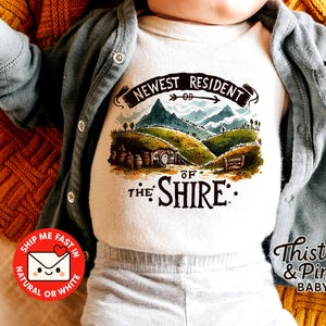 May include: White baby bodysuit featuring a landscape graphic with the text "NEWEST RESIDENT OF THE SHIRE". The design includes green hills, mountains, and a hobbit hole. The baby is wearing a grey cardigan.