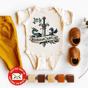 以下が含まれることがあります： 「Little Dungeon Crawler」の文字と剣とドラゴンのグラフィックが入ったオフホワイトのベビーロンパース。マスタード色のパンツ、茶色の革のベビーシューズ、木製のブロックも写っています。