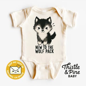 May include: Cream-colored baby onesie with a black and white wolf cub graphic. The text "NEW TO THE WOLF PACK" is printed below the wolf. The brand name "Thistle & Pine BABY" is also visible.