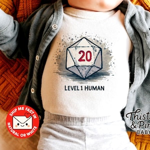 Op de afbeelding: Witte babybody met een afbeelding van een D20-dobbelsteen, met het nummer 20 in rood. De tekst "LEVEL 1 HUMAN" staat onder de dobbelsteen. De baby draagt een grijze cardigan en ligt op een oranje, getextureerde deken.