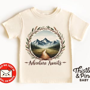 May include: A cream-coloured t-shirt with a watercolour design of a mountain landscape inside a circular wreath. The words "Adventure Awaits" are below the image. The brand name "Thistle & Pine BABY" is printed on the right side.