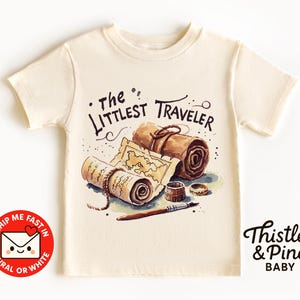 Peut inclure: T-shirt crème avec une illustration à l'aquarelle de cartes enroulées, d'un stylo et d'anneaux. Le texte "The Littlest Traveler" est au-dessus de l'illustration. Le t-shirt porte également le texte "Thistle & Pine BABY" et un cercle rouge avec du texte.