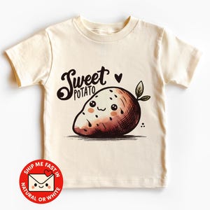 Könnte beinhalten: Cremefarbenes T-Shirt mit einem Cartoon-Design einer Süßkartoffel. Die Süßkartoffel hat ein lächelndes Gesicht und grüne Blätter. Die Worte "Sweet Potato" stehen in einer verspielten Schriftart über der Süßkartoffel. Ein kleines Herz ist darüber.
