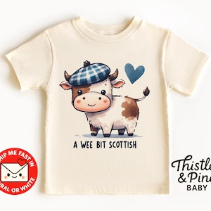 May include: Cream-colored baby t-shirt featuring a cartoon cow wearing a blue plaid beret and a blue heart. The text "A Wee Bit Scottish" is below the cow. The shirt also has the text "Thistle & Pine BABY".