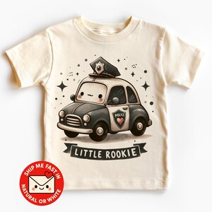 Könnte beinhalten: Cremefarbenes T-Shirt mit einem Cartoon-Polizeiauto mit lächelndem Gesicht, Polizeimütze und dem Wort "POLICE" auf der Tür. Der Text "LITTLE ROOKIE" befindet sich auf einem Banner darunter. Sterne und Glitzer im Hintergrund.