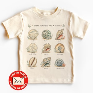 Op de afbeelding: Een crèmekleurig T-shirt met korte mouwen en een grafische print van negen verschillende schelpen in een raster. De tekst "EVERY SEASHELL HAS A STORY" staat erboven, waarbij elke schelp is gelabeld met de namen scallop, turbo, conch, sand dollar, murex, nautilus, cowrie, whelk en periwinkle.