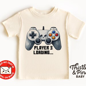 May include: A cream-coloured toddler t-shirt with a grey and black video game controller graphic. The text "PLAYER 3 LOADING..." is printed below the controller. The shirt has short sleeves and a round neckline.