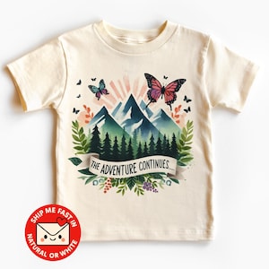 Könnte beinhalten: Cremefarbenes T-Shirt mit einem Aquarelldesign von Bergen, Schmetterlingen und Blättern. Die Grafik enthält den Text "THE ADVENTURE CONTINUES..." in einem Banner. Das Design beinhaltet Farbtöne von Blau, Grün, Rosa und Schwarz.