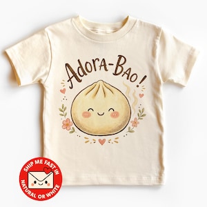 May include: A cream-coloured short-sleeve t-shirt featuring a central graphic of a smiling steamed bun with pink cheeks. Above the bun, the text reads Adora-Bao! in brown script, surrounded by small pink flowers, green leaves, and tiny red hearts.