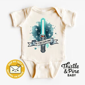 Könnte beinhalten: Cremefarbener Baby-Body mit einem Aquarelldesign eines blauen Lichtschwerts, Sternen und einem Banner mit der Aufschrift "THE CHOSEN ONE". Der Body hat kurze Ärmel und einen Wickelkragen. Der Markenname "Thistle & Pine BABY" ist unten rechts aufgedruckt.