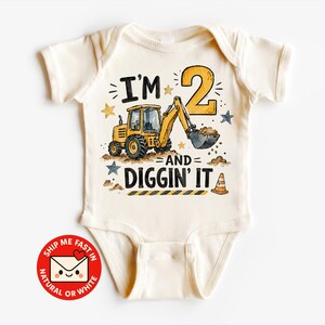 Könnte beinhalten: Ein cremefarbener Kurzarm-Babybody mit einer Grafik eines gelben Baggers, einer großen Zahl 2 und dem Text I'M 2 AND DIGGIN' IT. Das Design enthält kleine Sterne, Erdhaufen und einen Verkehrskegel in Gelb-, Blau- und Brauntönen.