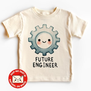 Könnte beinhalten: Cremefarbenes T-Shirt mit einem lächelnden Zahnrad-Motiv in Blau und Grau. Der Text "FUTURE ENGINEER" ist darunter gedruckt. Ein roter und weißer Aufkleber mit Text ist unten links sichtbar.