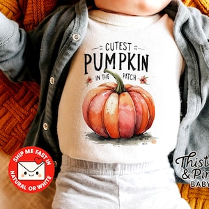 Könnte beinhalten: Ein weißer Baby-Body mit dem Text "Cutest Pumpkin in the Patch" und einer Kürbisgrafik im Aquarellstil. Das Baby trägt eine graue Strickjacke und liegt auf einer gelb-orangenen Strickdecke. Der Body ist für Babys.
