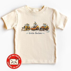 May include: A cream-coloured short-sleeve t-shirt featuring a graphic of three yellow construction vehicles, including a bulldozer, a road roller, and a front loader. The text "Little Builder" is printed in dark blue below the vehicles.