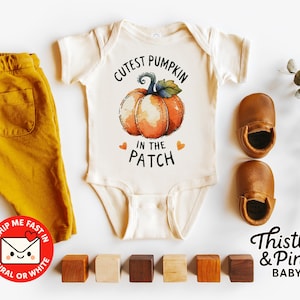 Könnte beinhalten: Ein cremefarbener Baby-Body mit einem Kürbis-Motiv und dem Text "Cutest Pumpkin in the Patch" wird mit senffarbenen Hosen, braunen Baby-Lederschuhen und Holzblöcken gezeigt. Ein Zweig Grünzeug ist ebenfalls vorhanden.