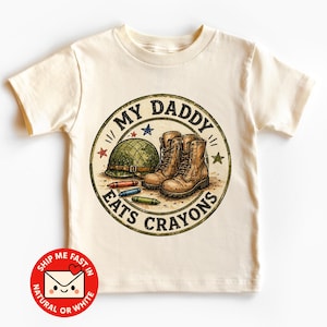 Könnte beinhalten: Ein cremefarbenes Kurzarm-T-Shirt mit einer kreisförmigen Grafik, die einen grünen Militärhelm, braune Kampfstiefel und drei Buntstifte zeigt. Der Text MY DADDY EATS CRAYONS ist mit kleinen roten, blauen und grünen Sternen im Vintage-Stil verziert.