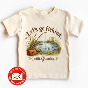 Könnte beinhalten: Ein cremefarbenes Kurzarm-T-Shirt mit einer grafischen Illustration einer Angelszene an einem Teich, inklusive Angelrute, Angelkasten und Seerosen. Der Text "Let's go fishing with Grandpa" ist in brauner Schreib- und Serifenschrift gedruckt.