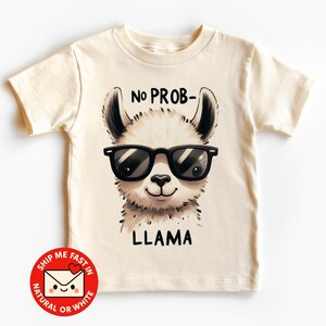 Könnte beinhalten: Cremefarbenes T-Shirt mit einem Cartoon-Lama, das eine schwarze Sonnenbrille trägt. Der Text "NO PROB-" steht über dem Kopf des Lamas und "LLAMA" darunter. Ein rot-weißer Aufkleber mit Text befindet sich unten links.