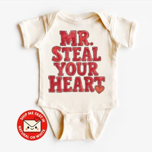 Könnte beinhalten: Ein cremefarbener Kurzarm-Babybody mit dem roten Schriftzug MR. STEAL YOUR HEART und einem kleinen roten Herzsymbol am Ende. Die Schrift hat eine texturierte Aquarell-Optik. Ein kreisförmiges rotes Abzeichen mit einem Briefumschlag-Symbol ist zu sehen.