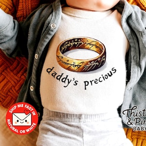 Pode incluir: Um body branco para bebé com um desenho de anel dourado e as palavras "daddy's precious" em preto. O bebé está a usar um cardigan cinzento e está deitado numa manta de malha amarela e laranja. O body é da Thistle & Pine Baby.