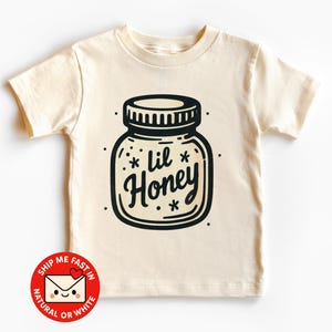 Peut inclure: T-shirt crème pour enfant avec un dessin noir et blanc d'un pot de miel et l'inscription "lil Honey". Le t-shirt à manches courtes a un col rond. Le motif comprend de petits accents d'étoiles.