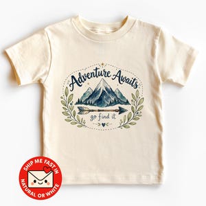May include: A cream-coloured short-sleeve t-shirt featuring a graphic print of blue mountains, a horizontal arrow, and green leafy branches. The text reads Adventure Awaits and go find it in a dark blue script font, surrounded by small stars and dots.