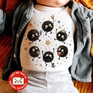 May include: A white short-sleeved bodysuit featuring a graphic print of five round, fuzzy black soot sprites with large eyes and thin limbs. The sprites are arranged in a circle, interacting with small, colorful stars in shades of yellow, blue, and orange.