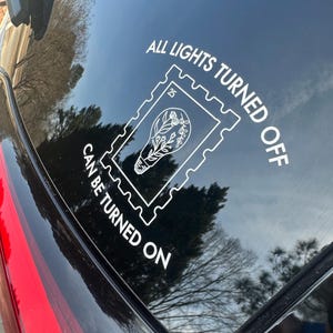Puede incluir: Una calcomanía blanca en la ventana de un coche negro con el texto "ALL LIGHTS TURNED OFF CAN BE TURNED ON". La calcomanía presenta un diseño de bombilla con elementos florales dentro de un marco similar a un sello postal.
