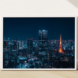May include: A framed print of a nighttime cityscape. The image features a vibrant, illuminated skyline with tall buildings and a prominent orange tower. The buildings are lit with blue and white lights against a dark blue sky.