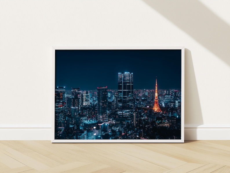May include: A framed print of a nighttime cityscape. The image features a vibrant, illuminated skyline with tall buildings and a prominent orange tower. The buildings are lit with blue and white lights against a dark blue sky.