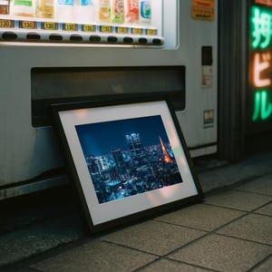 May include: A framed photograph of a nighttime cityscape, featuring illuminated skyscrapers and a prominent red tower. The photograph is leaning against a vending machine on a tiled surface, with a neon sign in the background.