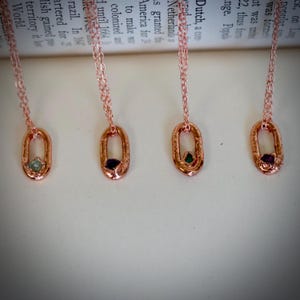 May include: Four rose gold necklaces with oval pendants, each featuring a different colored gemstone: green, purple, and two with a light green hue. The chains are delicate, and the pendants have a textured finish.